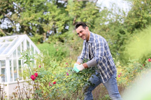 A man in a checkered shirt and jeans is tending to a garden in a residential outdoor space, surrounded by a variety of plants, including flowering bushes with pink blossoms and green foliage. He is smiling as he works among the garden beds, which are bordered by soil and low hedges. In the background, there is a white greenhouse structure with glass panels and a lush backdrop of tall trees and shrubs, indicating a well-maintained, landscaped garden. The garden surface features a mix of soil patches and grass, with some paved pathways visible. The weather appears bright and clear, suggesting a pleasant, sunny day suitable for outdoor gardening activities, aligning with professional gardening services such as those provided by Lawn Mowing Whitechapel in the local area.