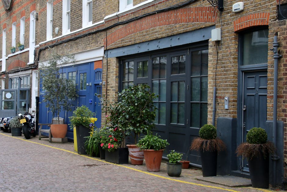 A narrow cobblestone street in Tower Hamlets lined with row houses featuring brick facades and large window panes. The front yards of these properties are landscaped with an array of potted plants, fl