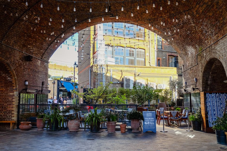 A view beneath a large brick brick archway showcasing a courtyard with a variety of potted plants and shrubs, including leafy green plants in terracotta and black pots arranged along the edge of the p