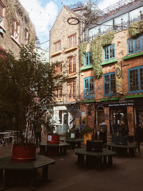 A view beneath a large brick brick archway showcasing a courtyard with a variety of potted plants and shrubs, including leafy green plants in terracotta and black pots arranged along the edge of the p
